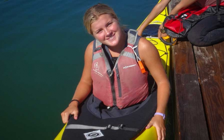 A young person wearing a life jacket sits in a kayak beside a dock and smiles at the camera